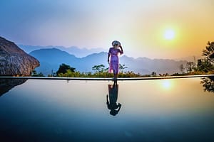 luxury travel on a budget, woman standing on a ledge with a hat and a mountain in the background